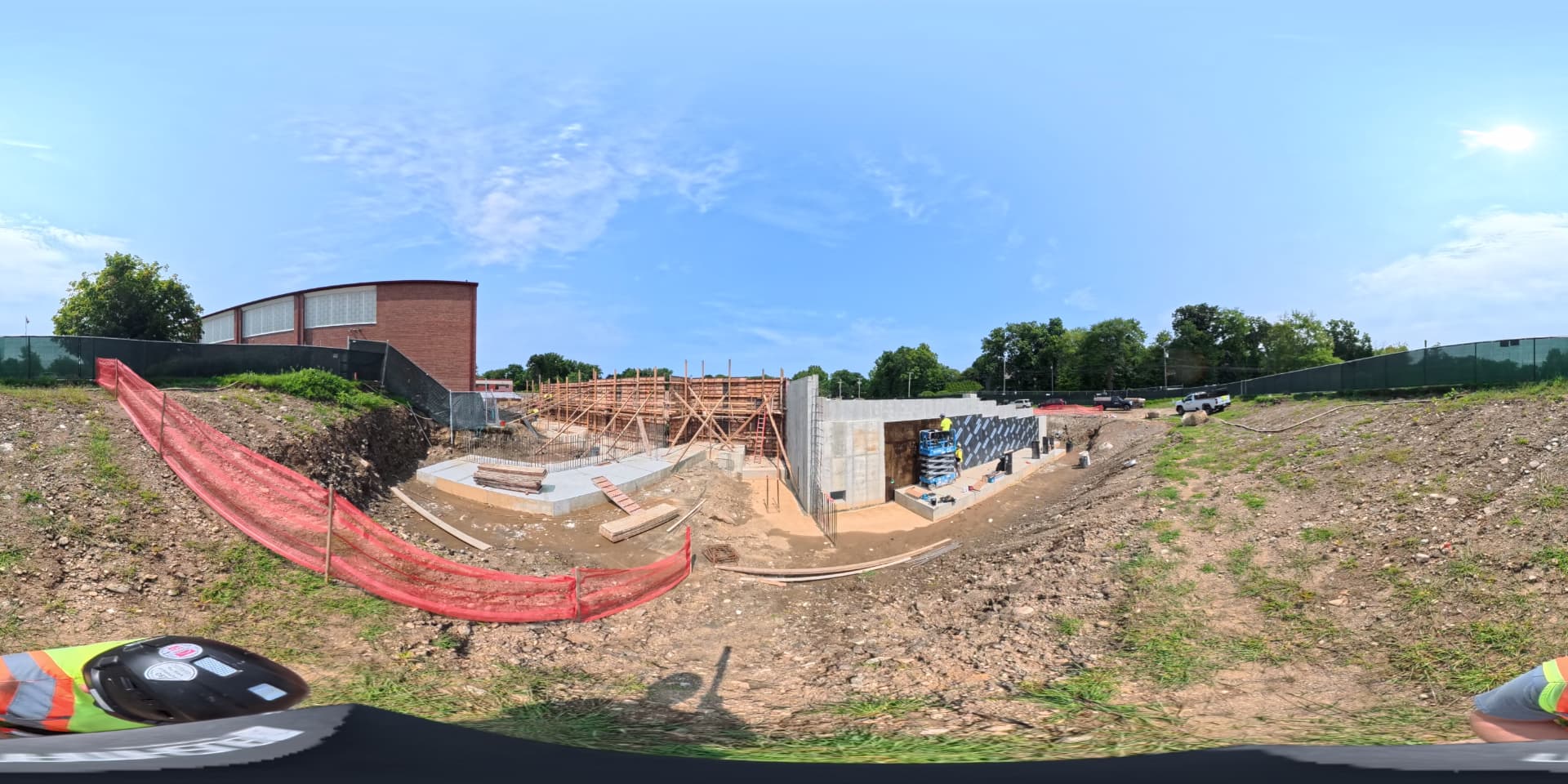August 2026 — Completed concrete foundation walls with waterproofing membrane applied, viewed from the excavated slope behind the entrance to the existing bus loop. 